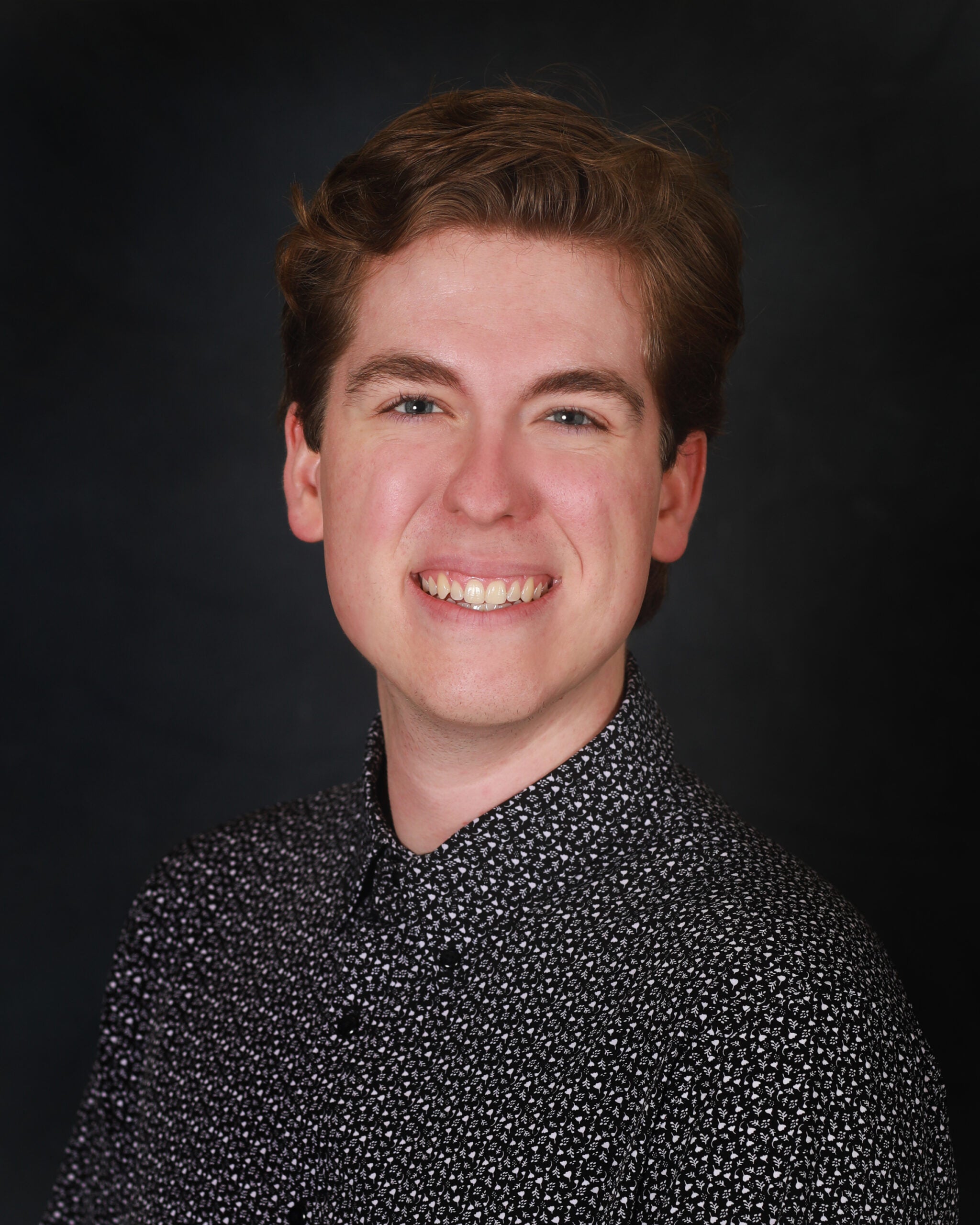 Headshot of a white man with brown hair and blue eyes, with a black collared shirt on a black backdrop.